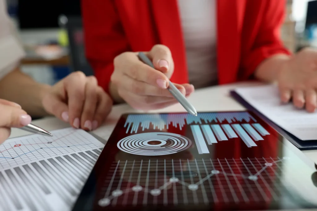 Professionals in red and white sit at a table discussing analytics and gesturing towards modern charts and graphs on a digital tablet.