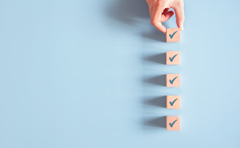 Five wooden blocks - each with a checkmark on it - are lined up against a light blue backdrop, a human hand setting in place the final top block.