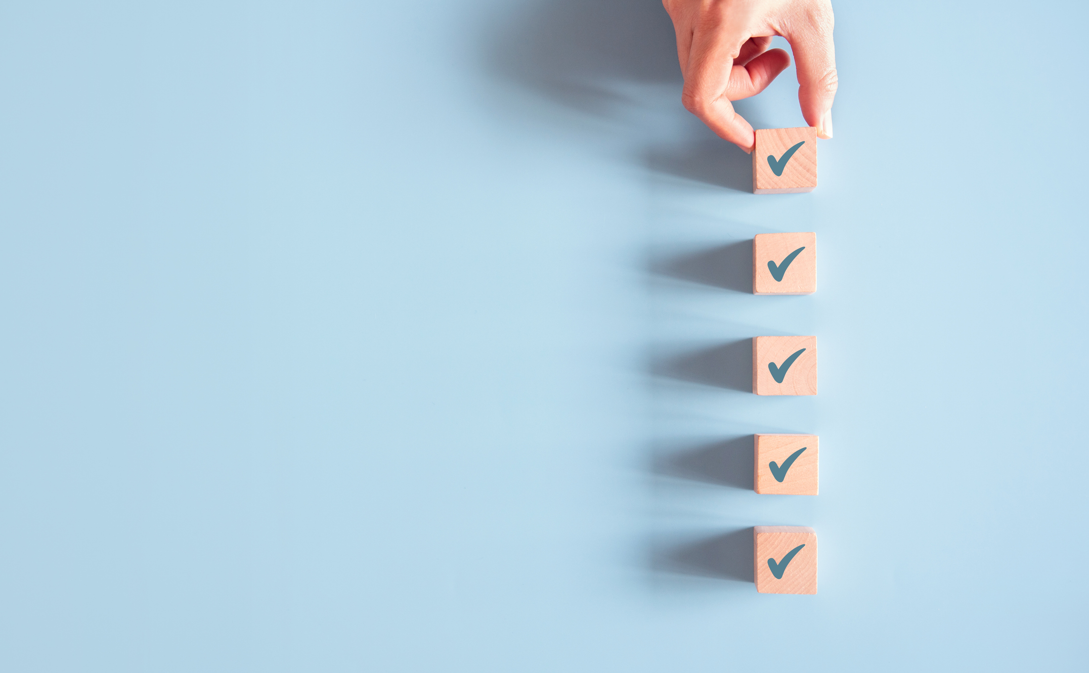 Five wooden blocks - each with a checkmark on it - are lined up against a light blue backdrop, a human hand setting in place the final top block.